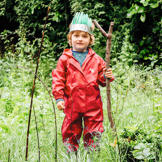 A child wearing an Originals Waterproof Puddle Suit Red stands in tall grass, a stick in hand, and a whimsical crown on their head.